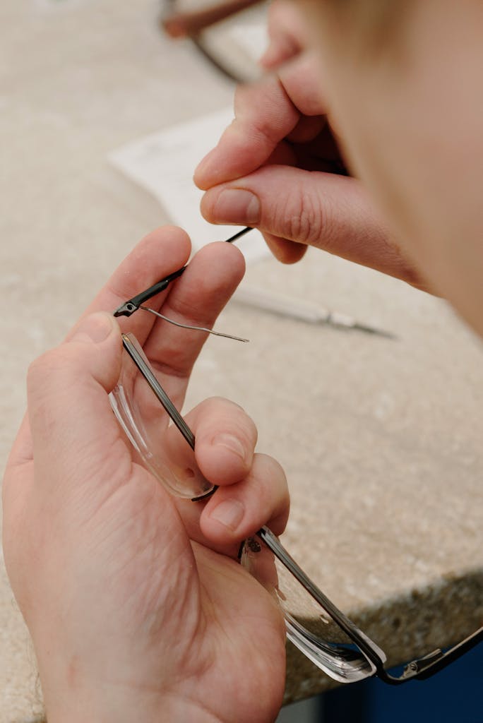 Unrecognizable male master repairing broken eyeglasses while sitting at table and attaching temple to hinge during work in professional workshop